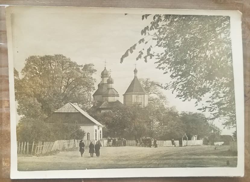 Дерев’яна церква з банями серед дерев, ліворуч хата під двосхилим дахом, дорогою йдуть люди
