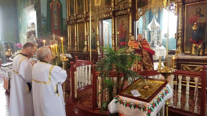 Священник у червоних ризах біля відчинених царських врат, поруч двоє вівтарників у білих стихарях; на аналої ікона з пальмовими гілками