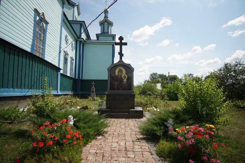 Доріжка з бруківки вздовж храму веде до гранітної стели з хрестом і образом Богородиці з Дитям, обабіч квіти
