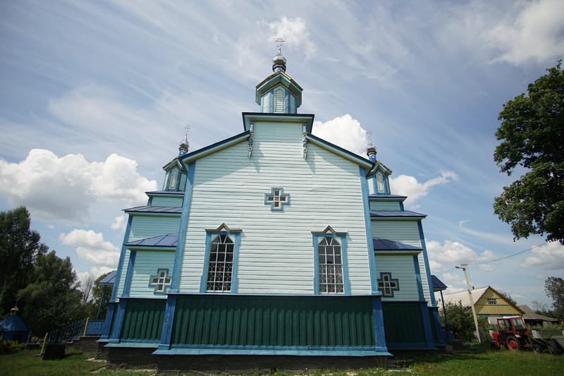 Фасад храму без входу: два високі вікна, хрестоподібні віконця, над дахом бані; праворуч сільська хата і червоний трактор