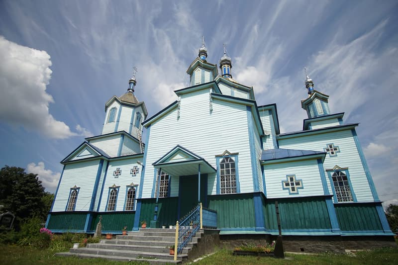 Блакитний дерев’яний храм із ганком і широкими сходами, над дахом бані на тлі неба з перистими хмарами