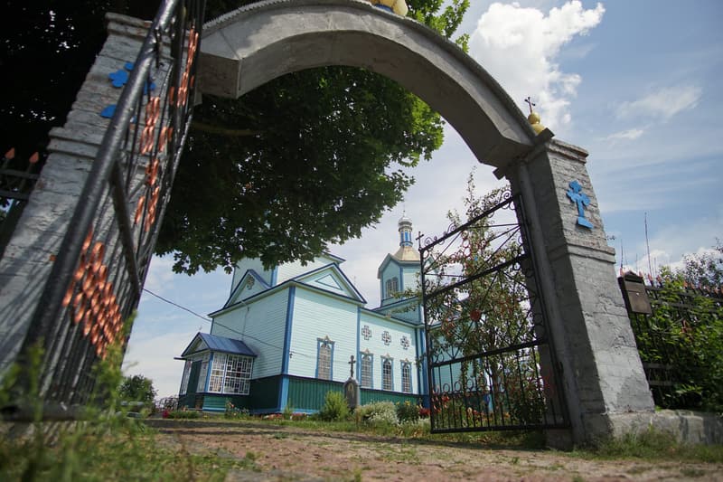 Храм у Рижанах крізь кам’яну арку брами з відчиненими кованими ворітьми
