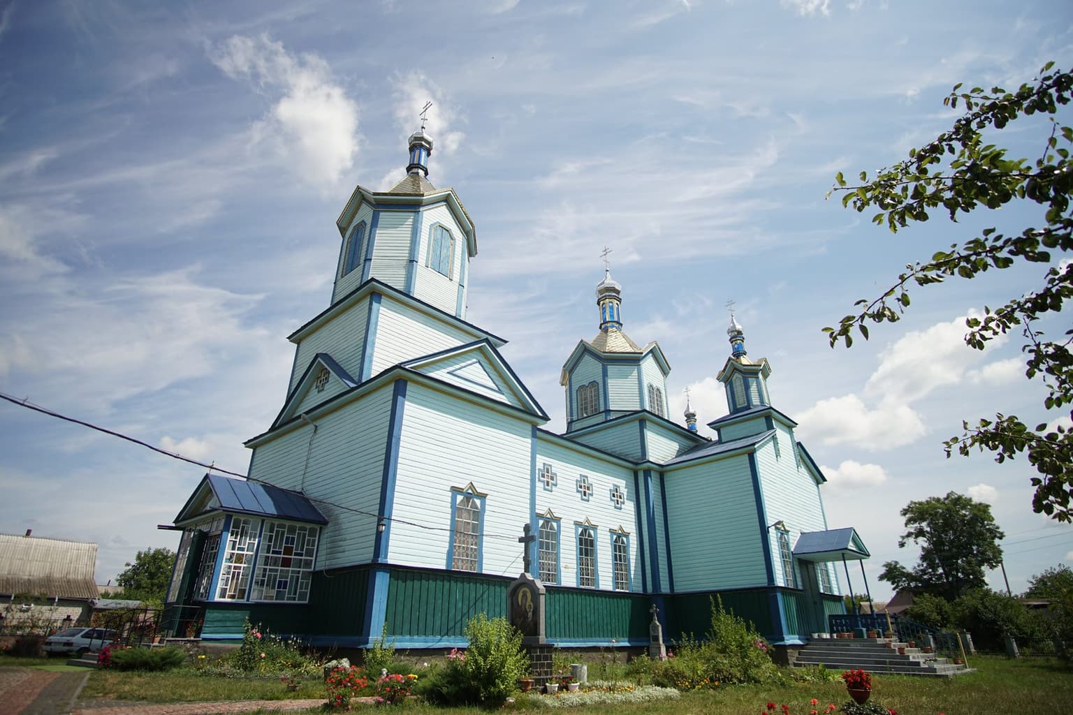 Дерев’яний храм Різдва Пресвятої Богородиці в Рижанах збоку: блакитні стіни з синім оздобленням, бані з хрестами, перед храмом квітник