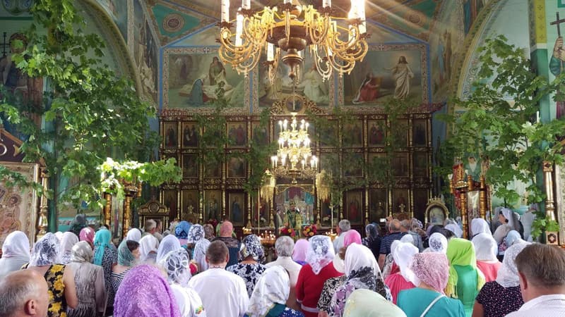 Парафіянки в світлих хустках на службі, храм прибраний зеленими гілками, над головами світяться люстри