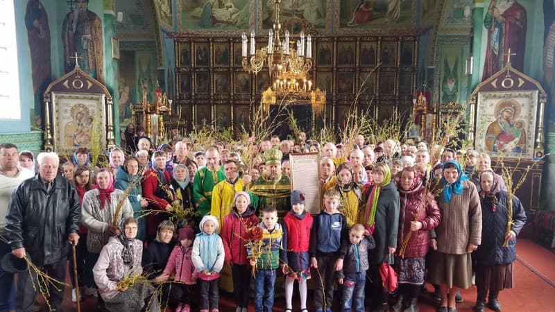 Парафіяни з дітьми й гілочками верби перед іконостасом, у центрі священник у зелених ризах і документ у рамці